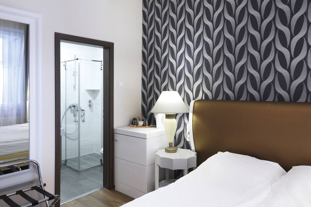 Ein Hotelzimmer mit Bett, Nachttisch mit Lampe, Duschbereich und Wand mit Muster.