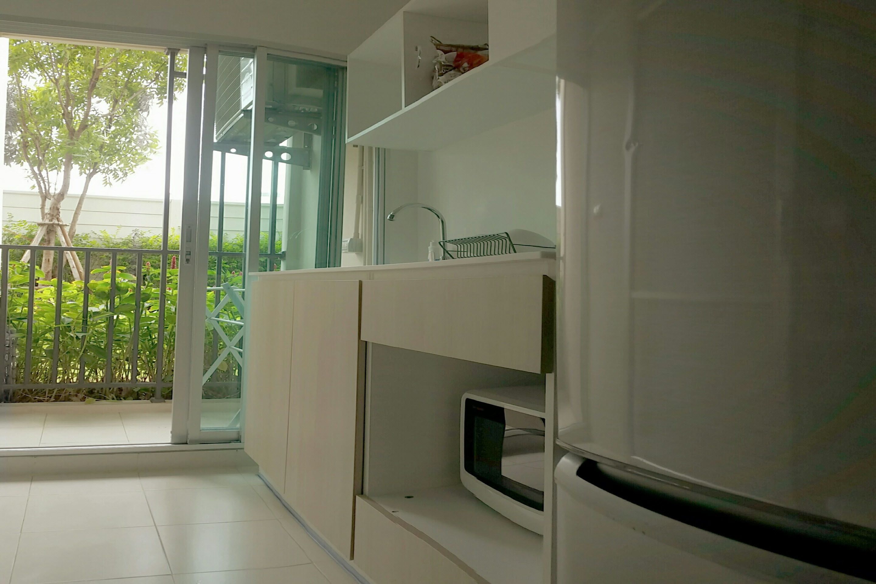 Une cuisine moderne avec des armoires blanches, un four à micro-ondes encastré et une porte-fenêtre coulissante donnant sur un balcon avec vue sur la verdure.