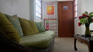 Un espace d’accueil avec un canapé en osier, une porte en bois et un panneau indiquant les équipements disponibles, tels que la climatisation, un ventilateur et de l’eau chaude.