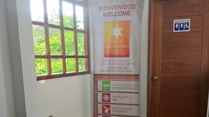 Une chambre d’hôtel avec un lit, un canapé, une fenêtre donnant sur un espace verdoyant, un panneau indiquant les équipements disponibles et une porte en bois.