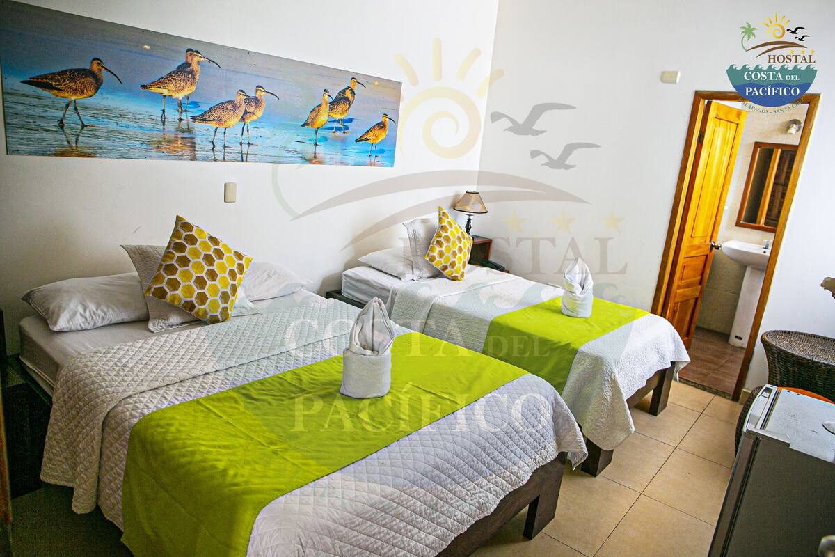 Een hotelkamer met twee bedden, elk met een groene sprei en een witte gebogen deken. Daarboven hangt een schilderij van vogels aan de muur, en rechts op de muur bevindt zich het logo van het hotel.