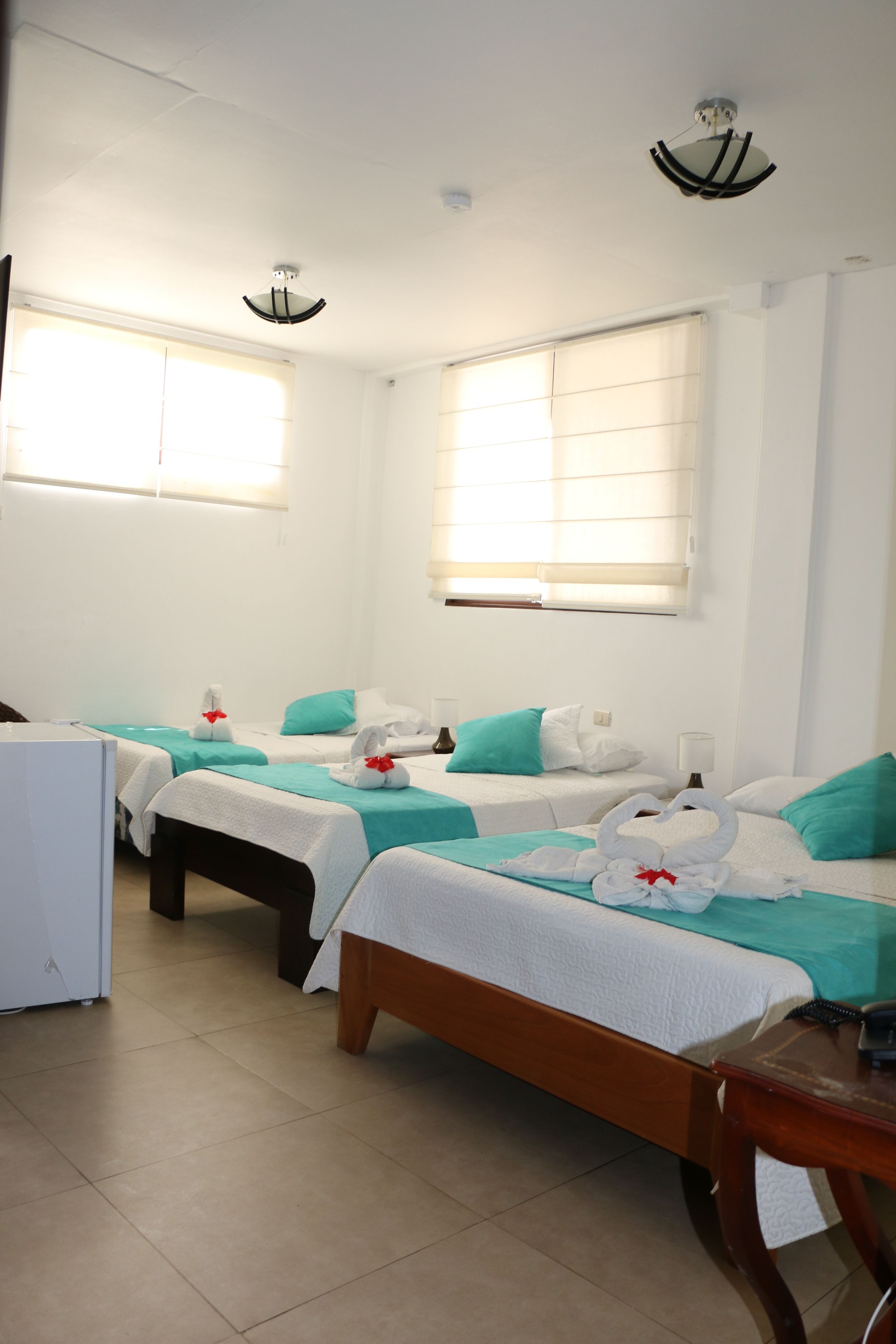 Habitación de hotel con dos camas, cada una con colcha color turquesa y ropa de cama blanca, una mesita de noche de madera, una pequeña mesa y un refrigerador blanco.