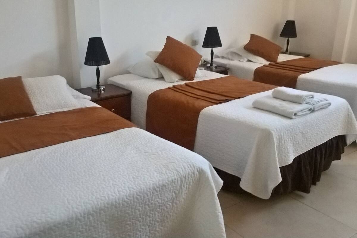 Een hotelkamer met vier bedden, elk met een witte spreidek en een bruin hoofdeinde. Op elk nachtkastje staan zwarte lampen.