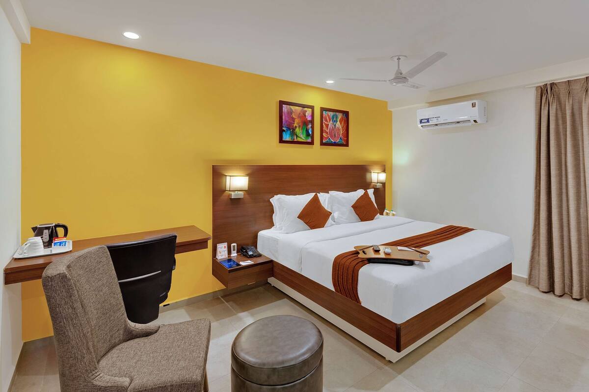 A hotel room with a bed, bedside tables, a chair, a desk, a coffee maker, a telephone, a remote control, a TV, an air conditioner, and a ceiling fan.