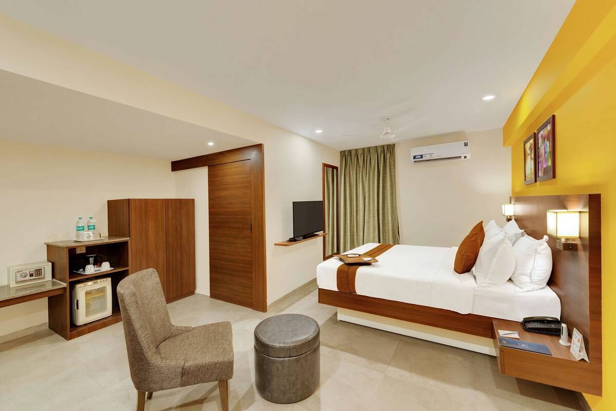 A hotel room with a bed, a desk, a chair, a television, a wardrobe, and a small round ottoman.