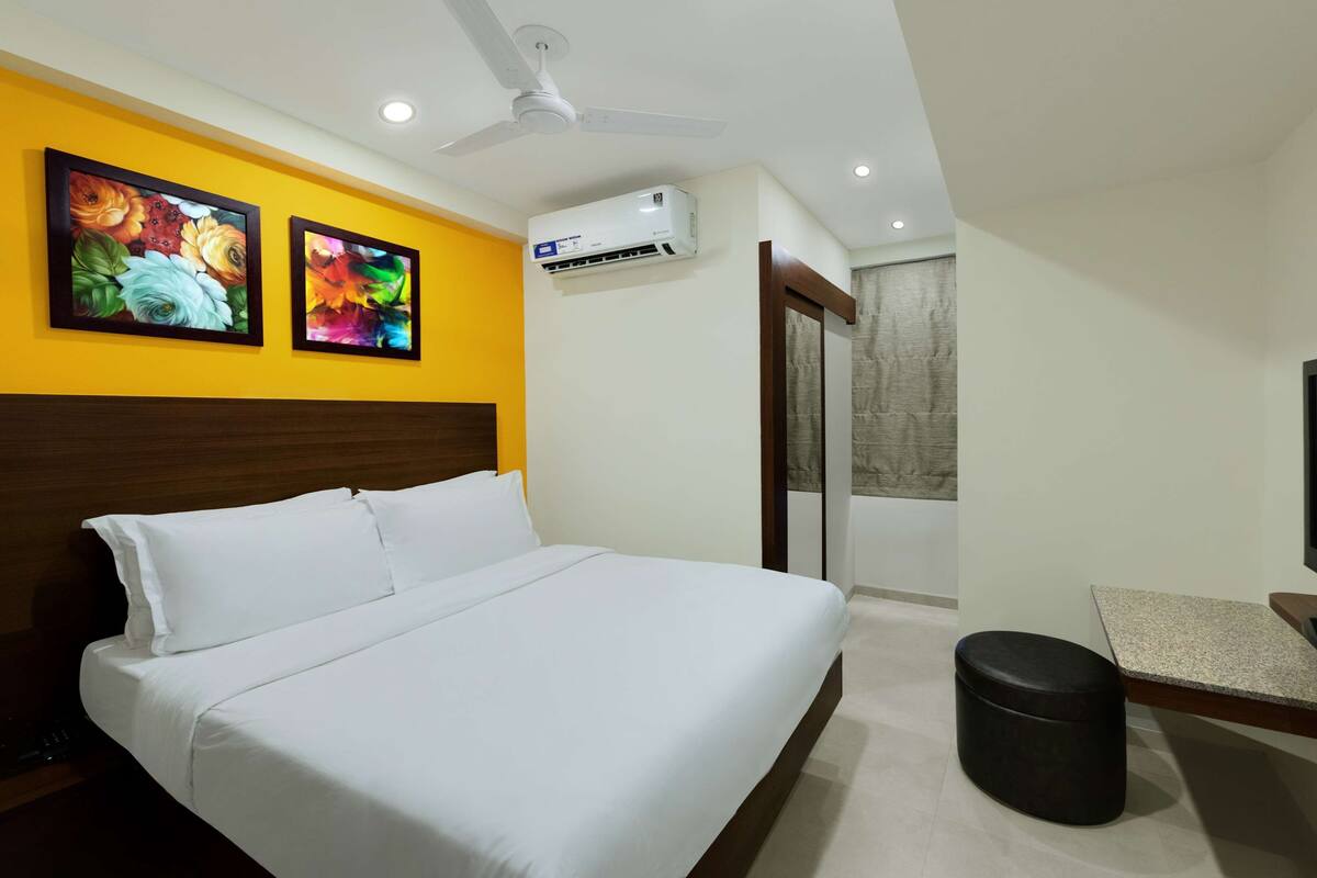 A hotel room with a bed, a ceiling fan, an air conditioning unit, and two paintings on the wall.