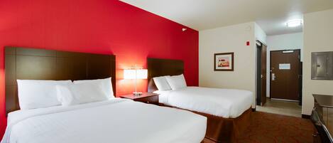 A hotel room with two beds, a red accent wall, a wooden headboard, a nightstand with a lamp, and a door leading to a bathroom.