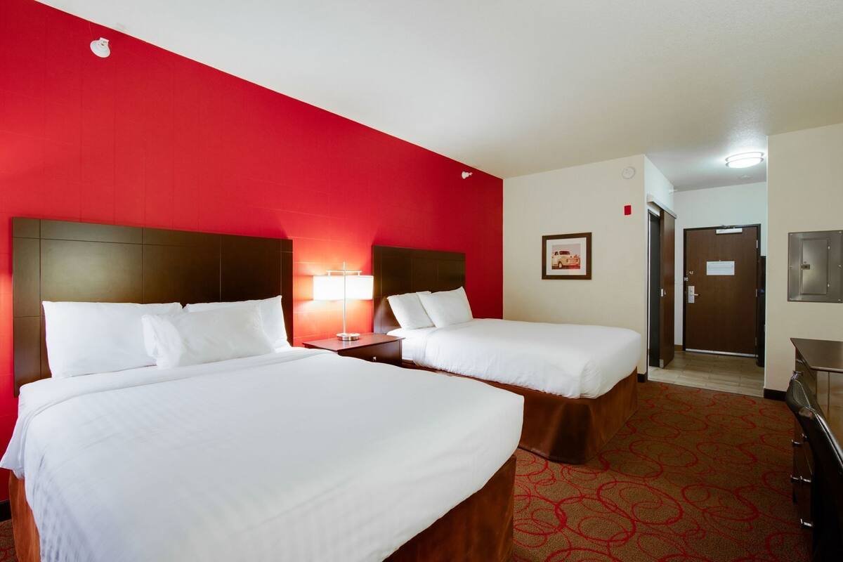 A hotel room with two beds, a red accent wall, a framed picture, and a door leading to a hallway.