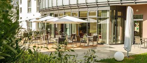 An outdoor seating area with tables and umbrellas, surrounded by glass windows and a building facade.