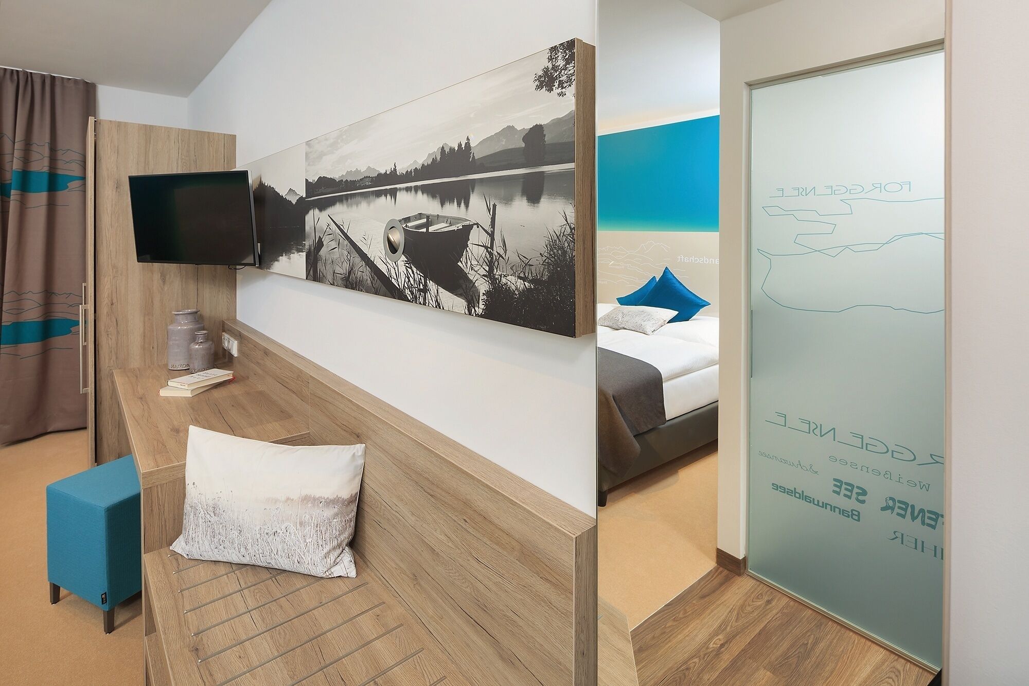 Quarto de hotel com cama, televisão, pintura grande, escrivaninha e divisória de vidro com texto.