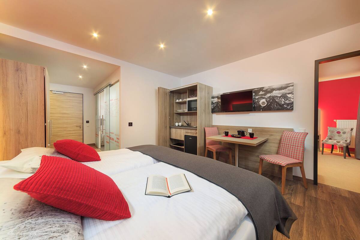 A modern hotel room with a large bed, a desk with a chair, a TV, and a red accent wall.