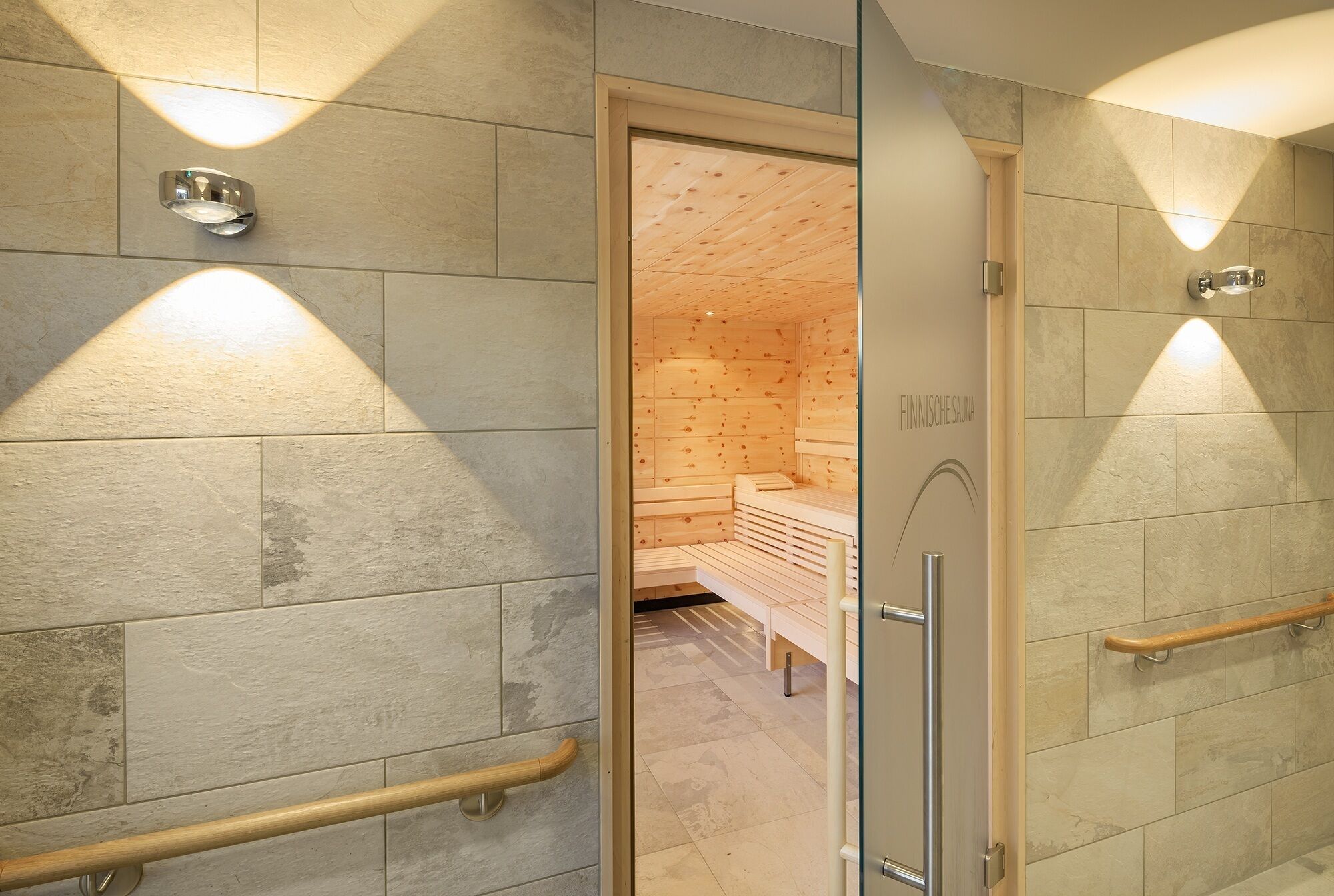 Un baño moderno con baldosas de piedra, bancos de madera y una puerta de cristal que da a un cuarto de sauna.