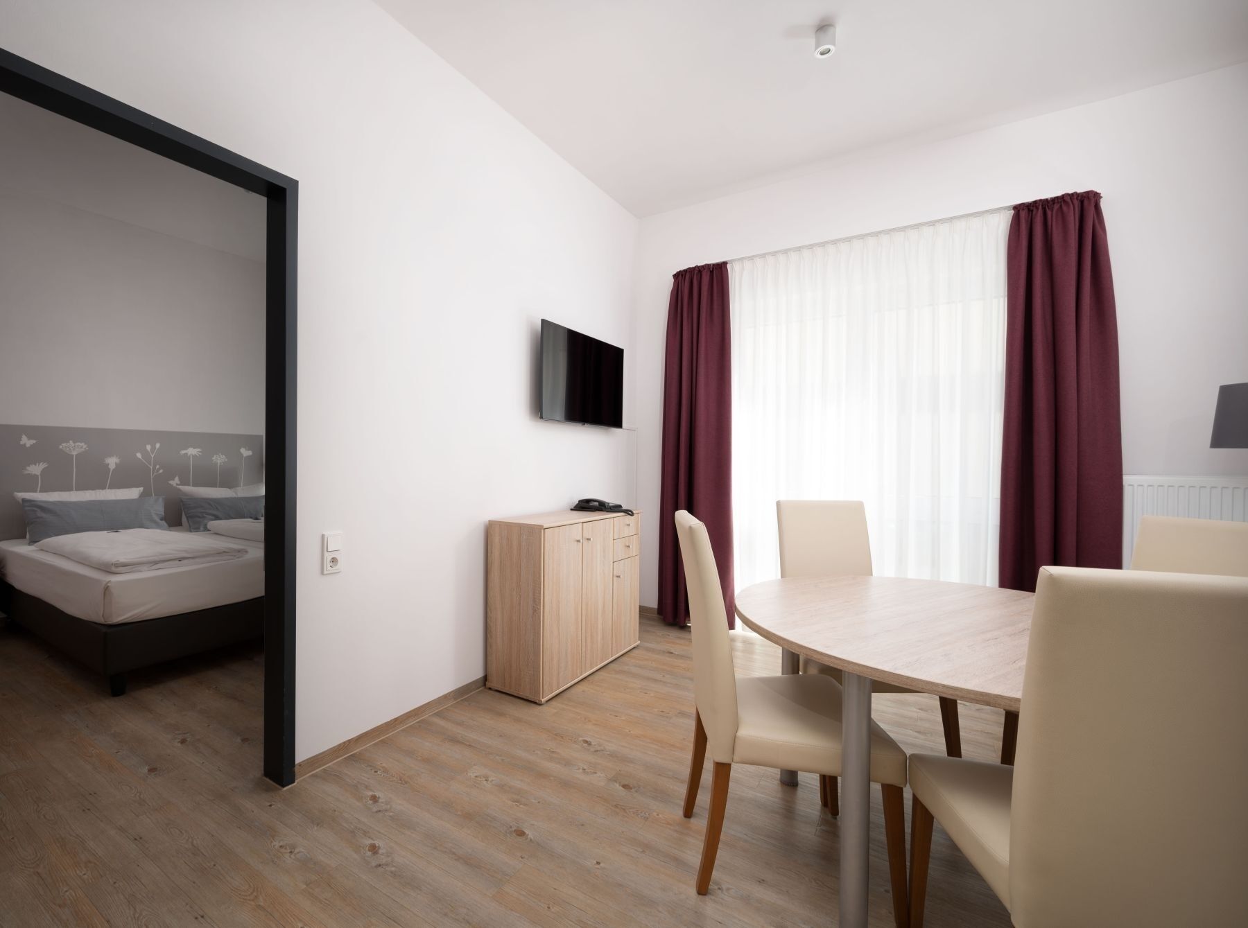 A modern hotel room with a bed, a wooden cabinet, a TV, and a dining area with chairs and a table.