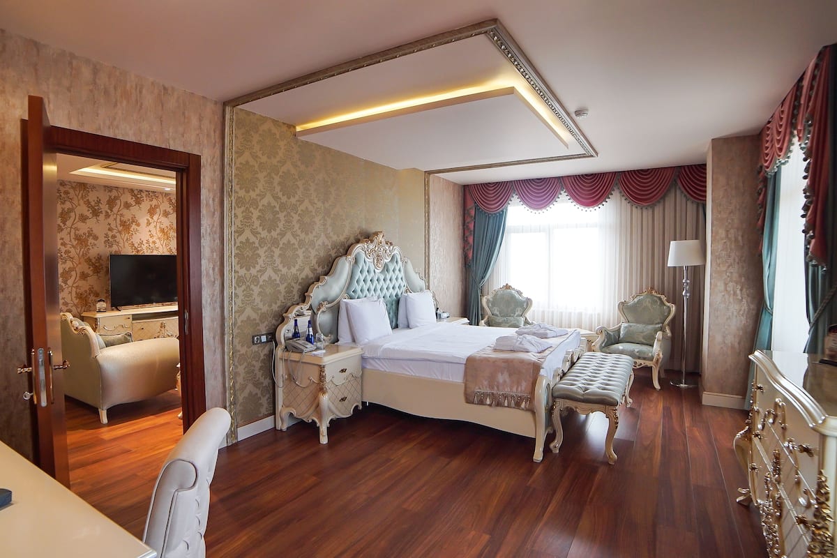 A spacious hotel room with a large bed, a sitting area with a chair and a table, a television, and a desk with a lamp.