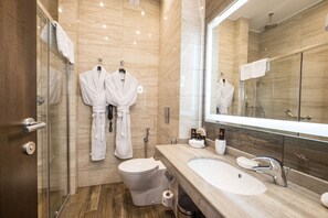 A modern bathroom with a glass shower enclosure, a toilet, a sink with a mirror, and two white robes hanging on a rack. - Tom River Plaza (Kemerovo)