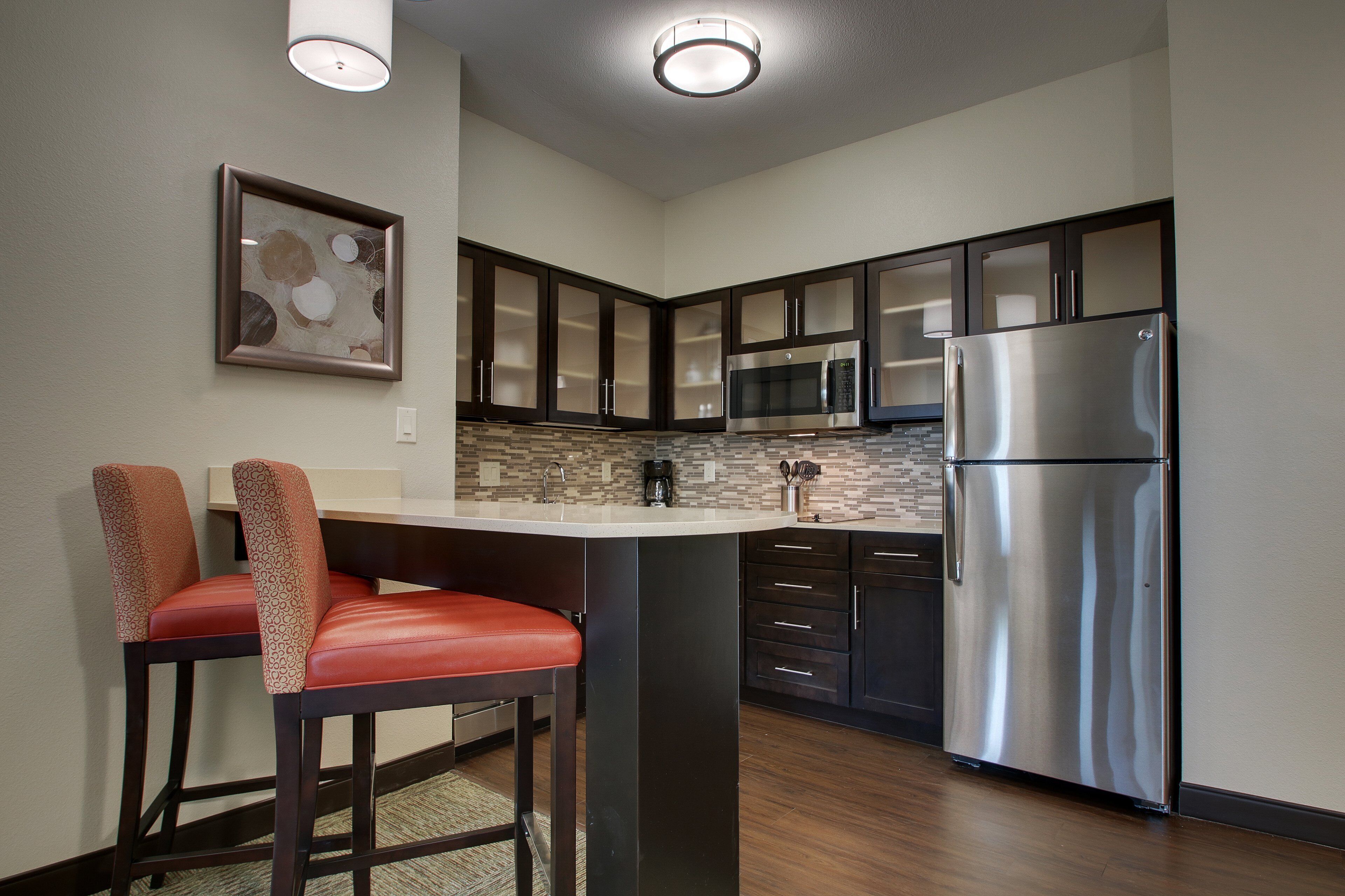 A modern kitchen with dark cabinets, a stainless steel refrigerator, and a bar area with two red chairs.