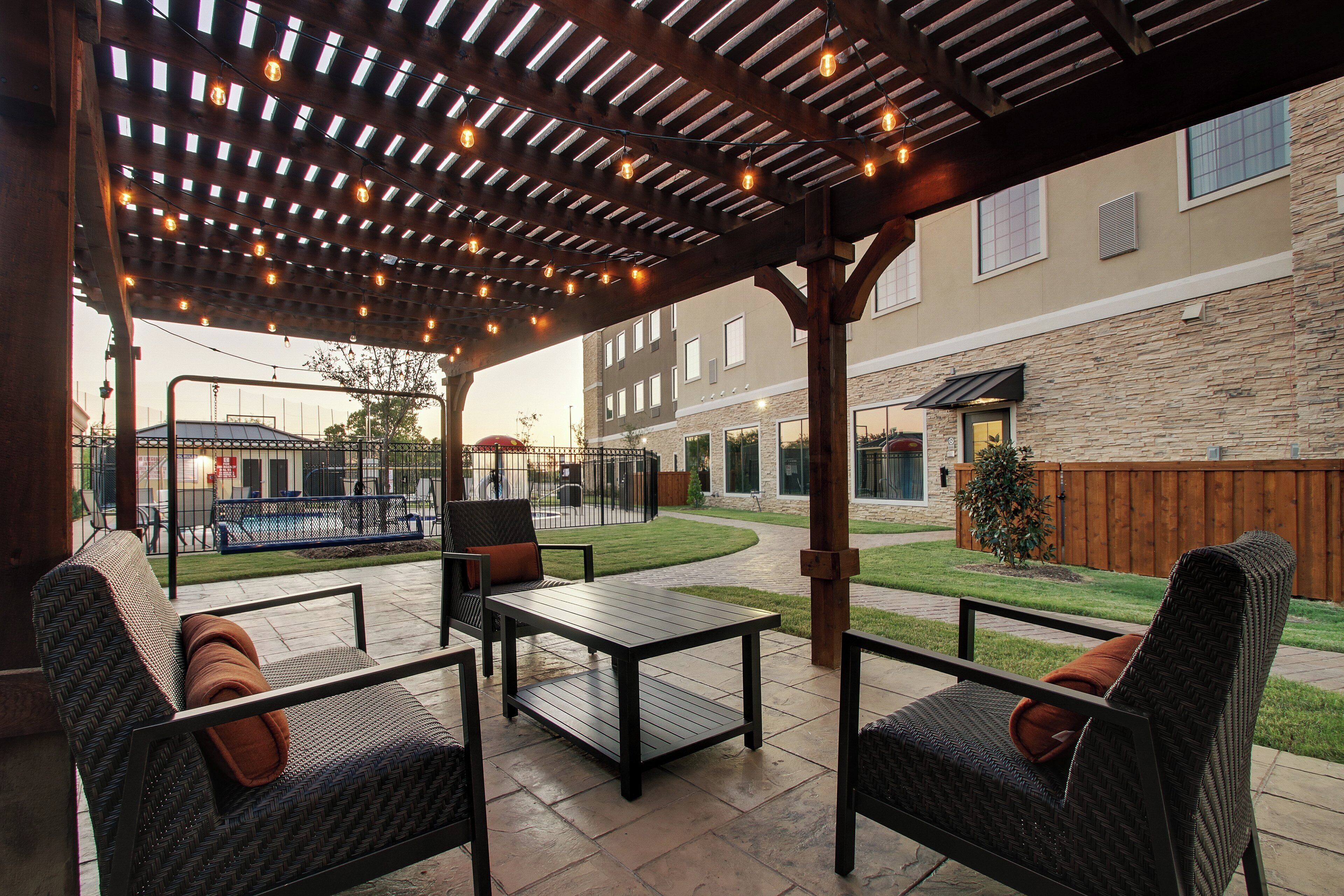 Un área de patio exterior con una pérgola, asientos y vistas a una piscina y edificios al fondo.