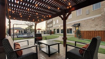 Un área de patio exterior con una pérgola, asientos y vistas a una piscina y edificios al fondo.
