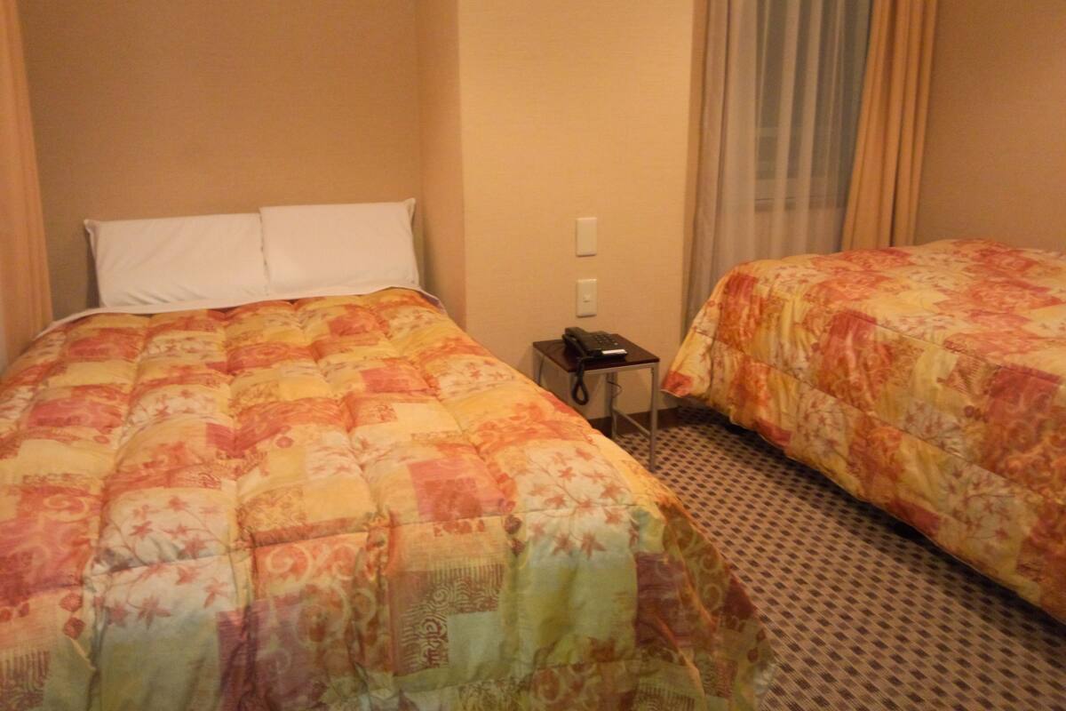 A hotel room with two beds, each with floral-patterned bedding, a small bedside table with a phone, and a window with curtains.