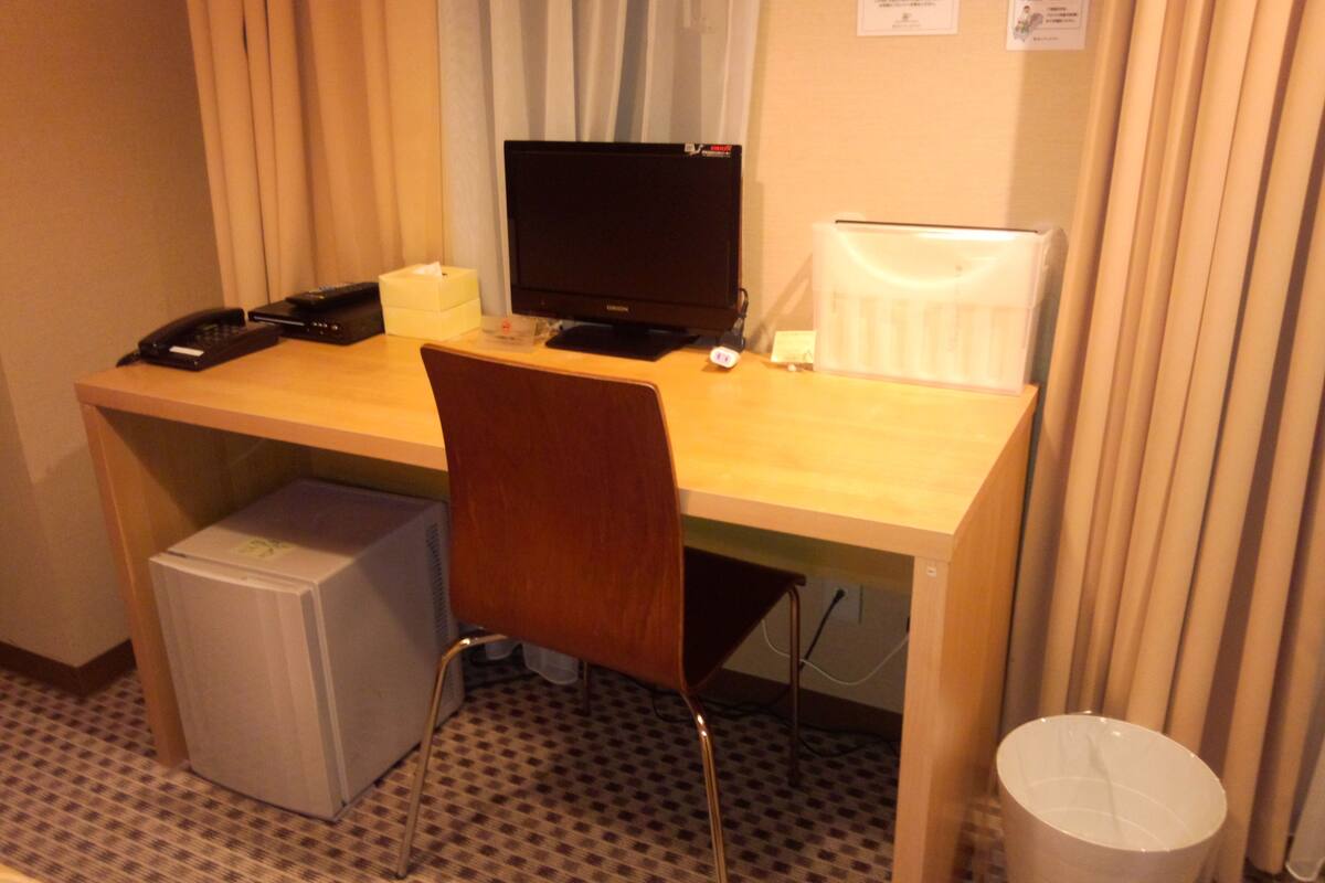 A hotel room with a wooden desk, a computer, a chair, a trash bin, and a small refrigerator.