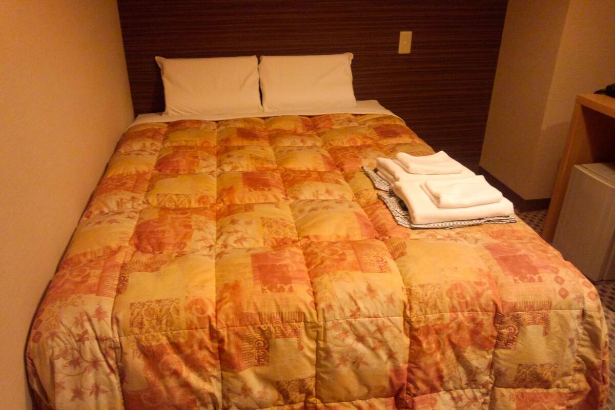 A neatly made bed with a patterned bedspread, two pillows, and folded towels on the side.