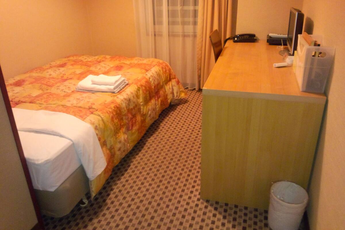 A hotel room with a bed, a desk, a computer, and a trash can.