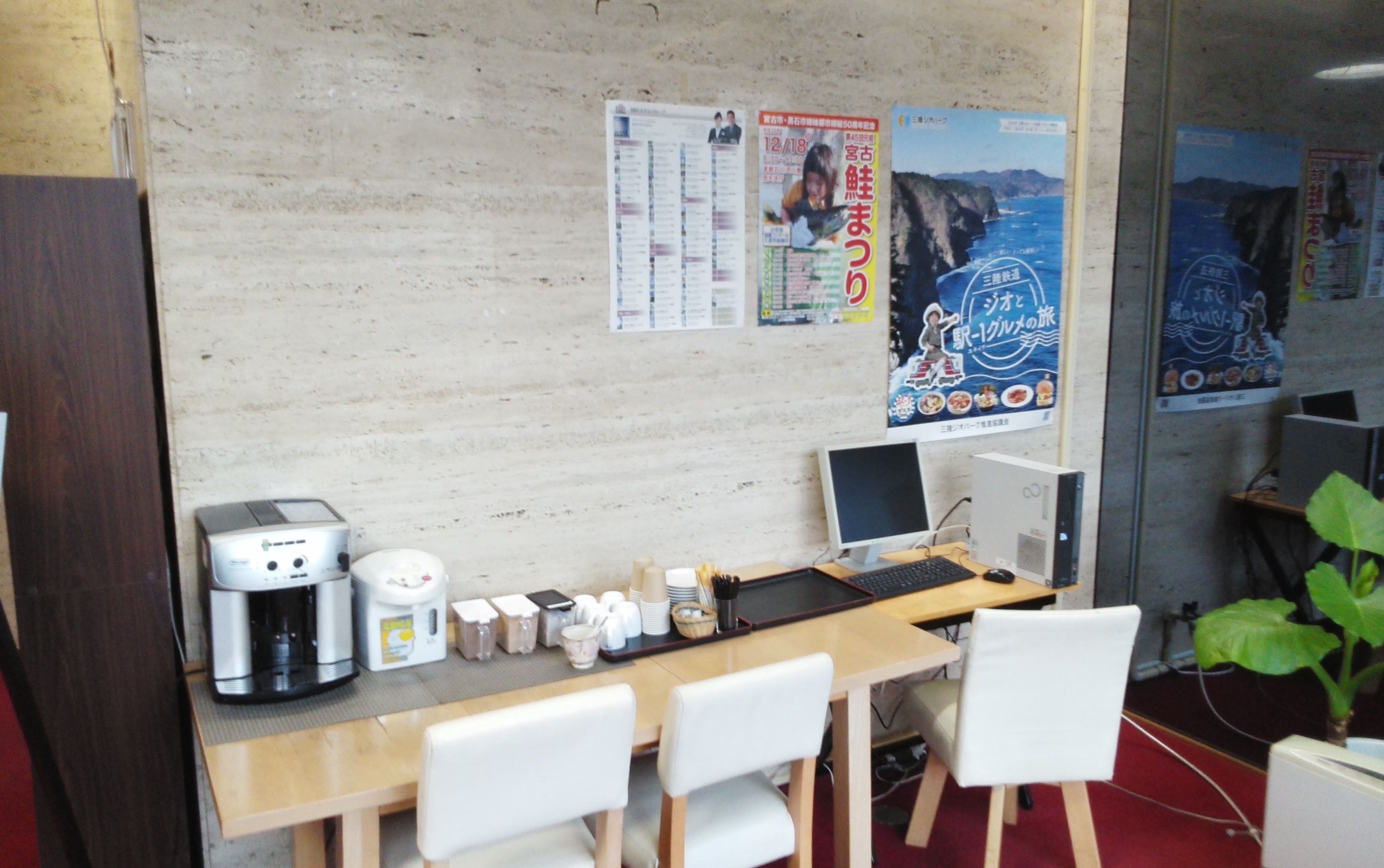 Ett litet kontor med ett skrivbord, en dator och en kaffebryggare. Det finns affischer på väggen och en växt i hörnet.