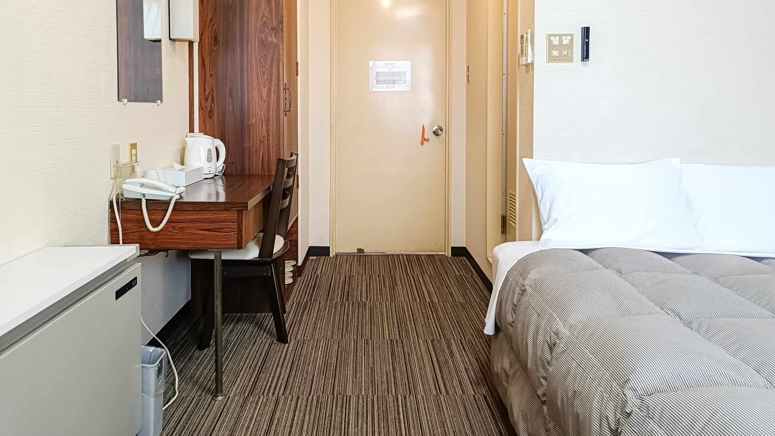 A hotel room with a bed, a desk with a telephone and a kettle, a chair, and a door with a sign.