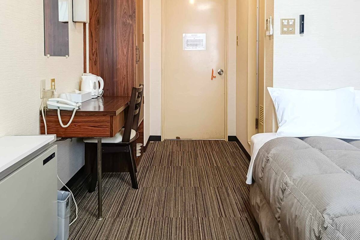 A hotel room with a bed, a desk with a telephone and a kettle, a chair, and a door with a sign.