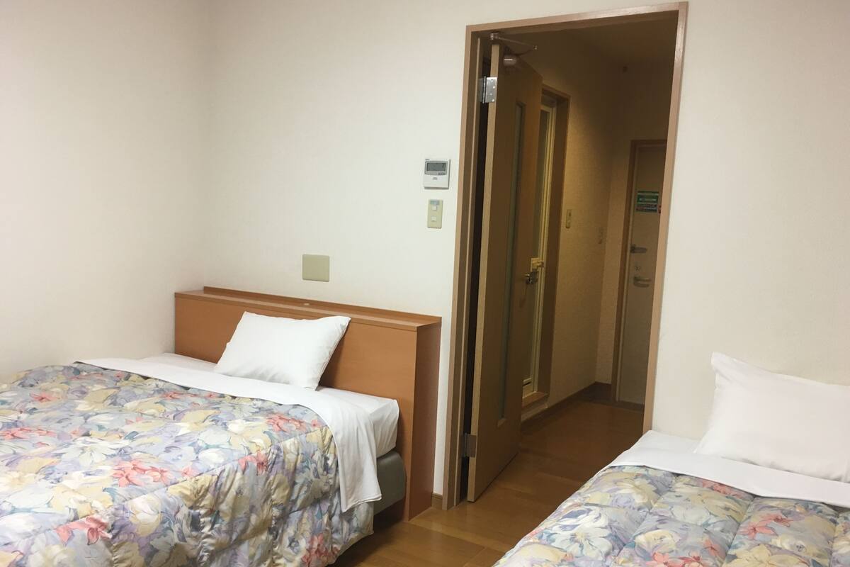 Ein Hotelzimmer mit zwei Einzelbetten, Holz-Kopfteilen, floralem Bettbezug, Holzboden, einer Tür zu einem Flur und einer kleinen Wanduhr.