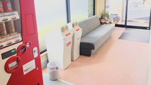 A vending machine with beverages, a Coca-Cola cooler, and a grey sofa in a well-lit indoor area with large windows. - Hotel Tajimi Hills My Room (Tajimi)