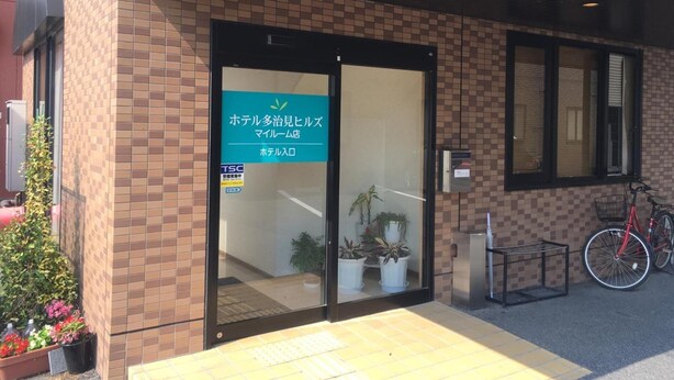 A hotel entrance with a sign in Japanese, a bicycle parked outside, and potted plants near the entrance. - Hotel Tajimi Hills My Room (Tajimi)