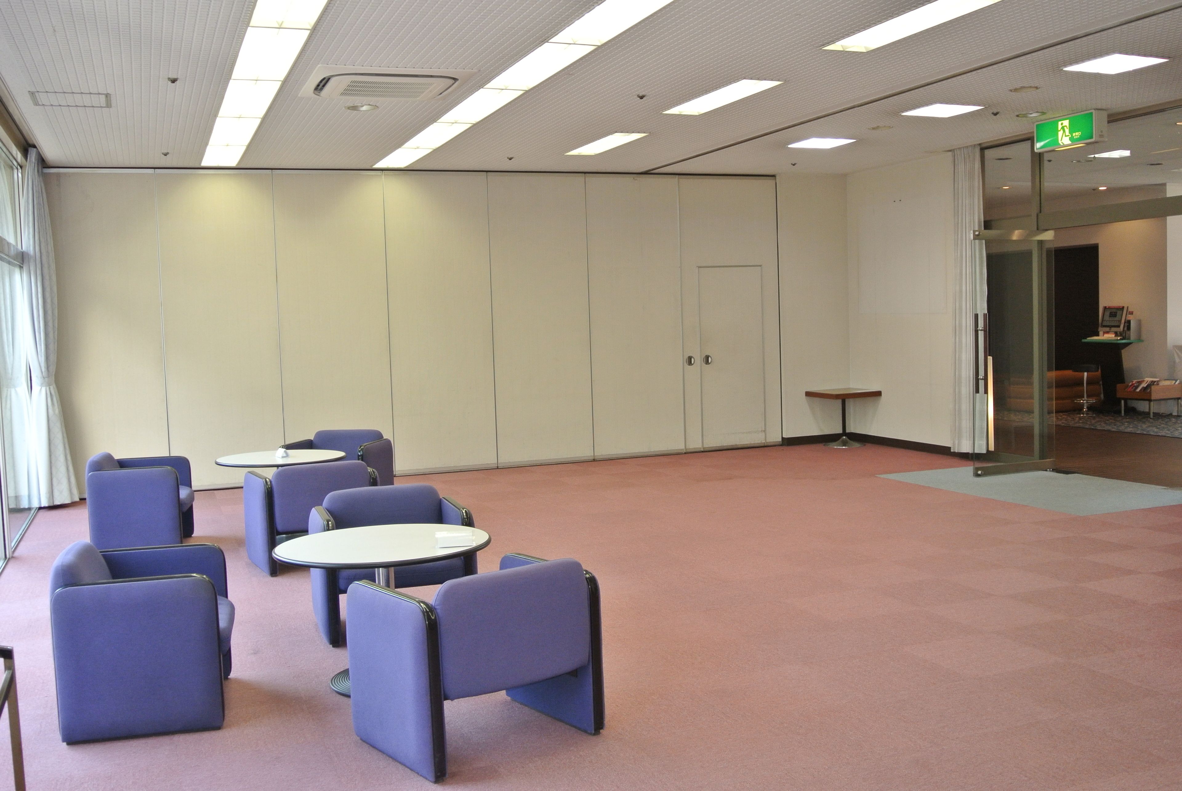 Un salón de conferencias con sillas y mesas azules, una puerta de cristal e iluminación empotrada en el techo.