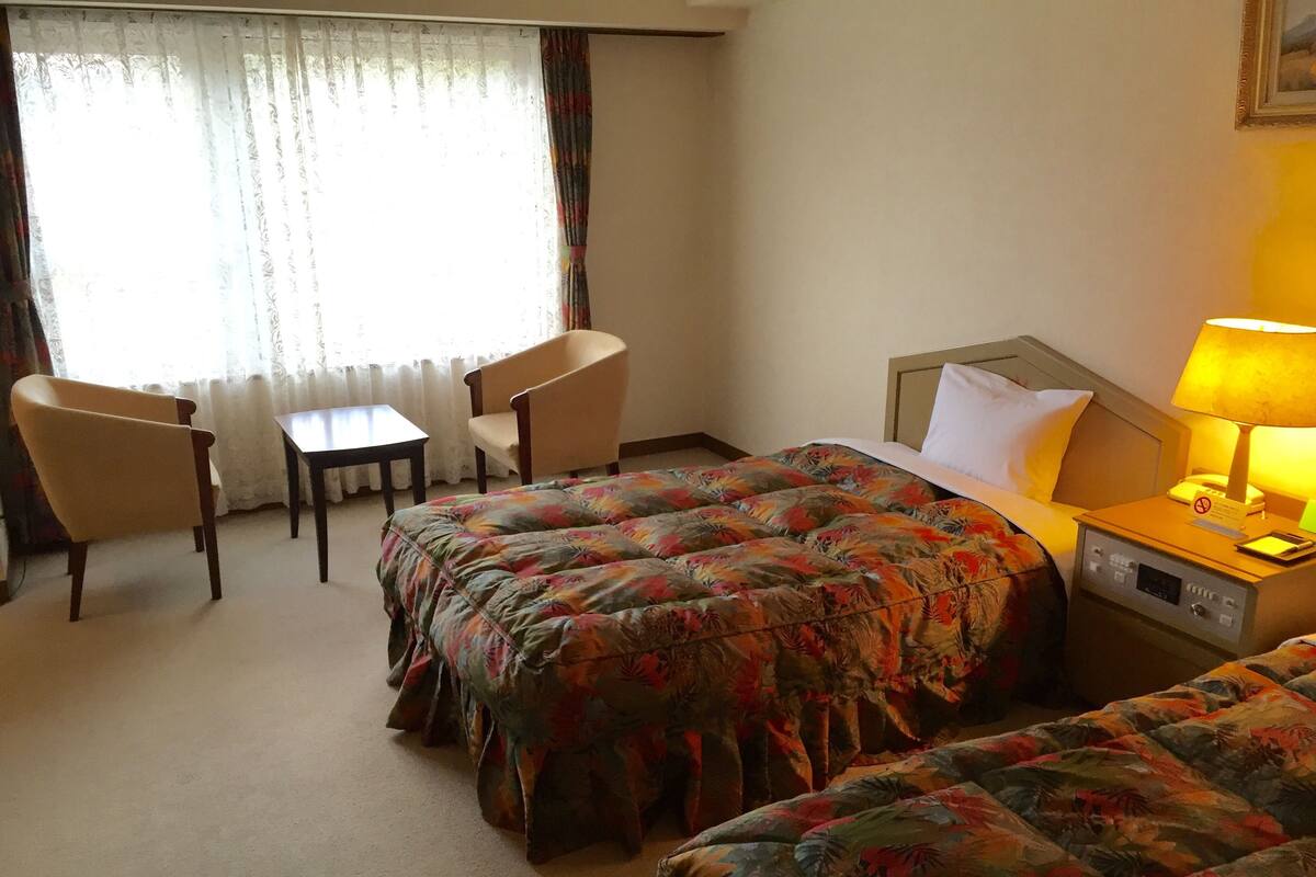 A hotel room with two beds, a chair, a table, and a lamp.