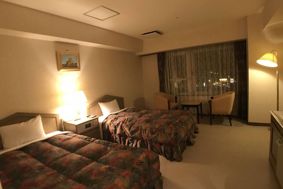 A hotel room with two beds, a desk, a chair, a lamp, and a window with curtains.