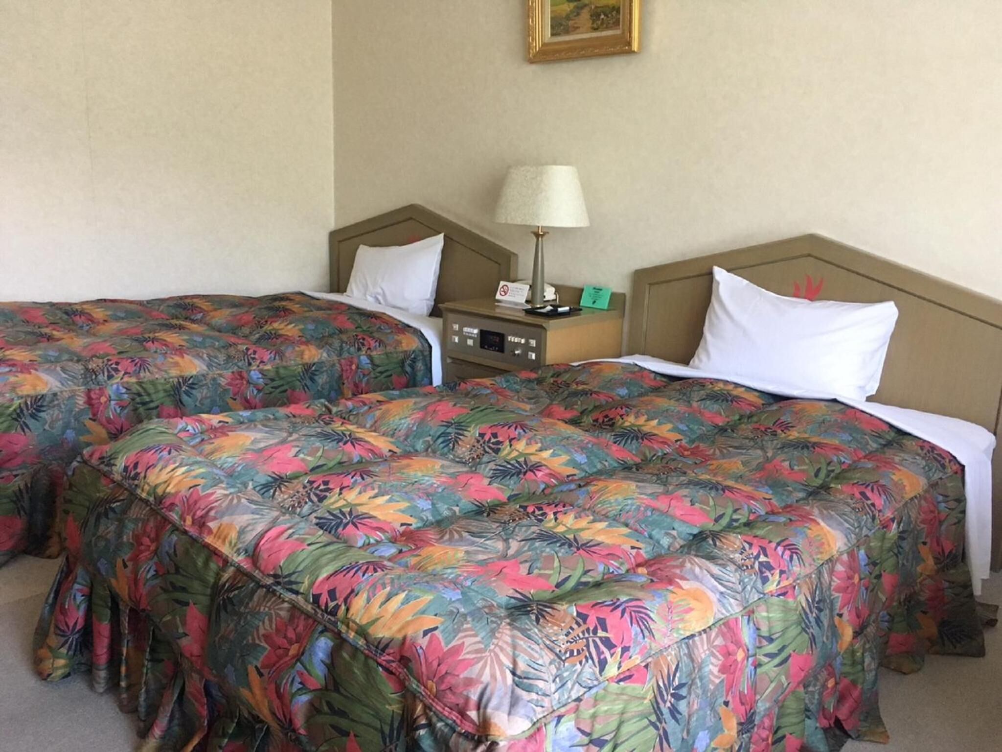 A hotel room with two beds, each with floral-patterned bedding, a bedside table with a lamp, and a small clock.