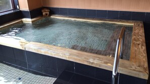 Un jacuzzi con borde de madera y barandilla metálica, rodeado de paredes de baldosas negras y un panel de madera en la pared trasera.