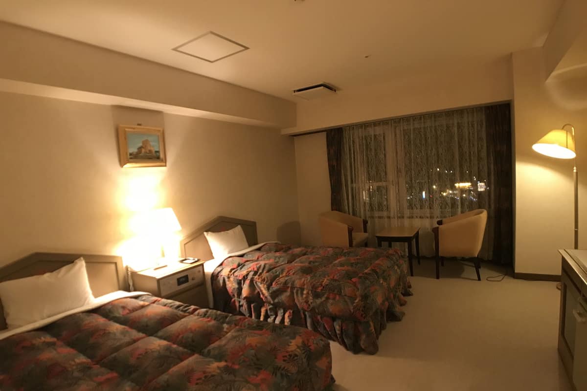 A hotel room with two beds, a desk, a chair, and a large window with curtains.