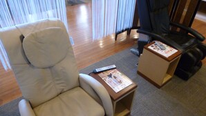 Une chambre d’hôtel avec un fauteuil de couleur beige, une petite table d’appoint en bois sur laquelle se trouve un magazine, et un fauteuil de bureau noir.