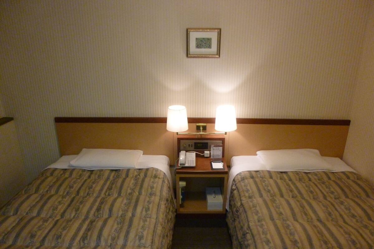 Hotelkamer met twee bedden, elk met een geborduurde spreideken, een nachtkastje met een lamp, een telefoon en een klein tafeltje met een menukaart.