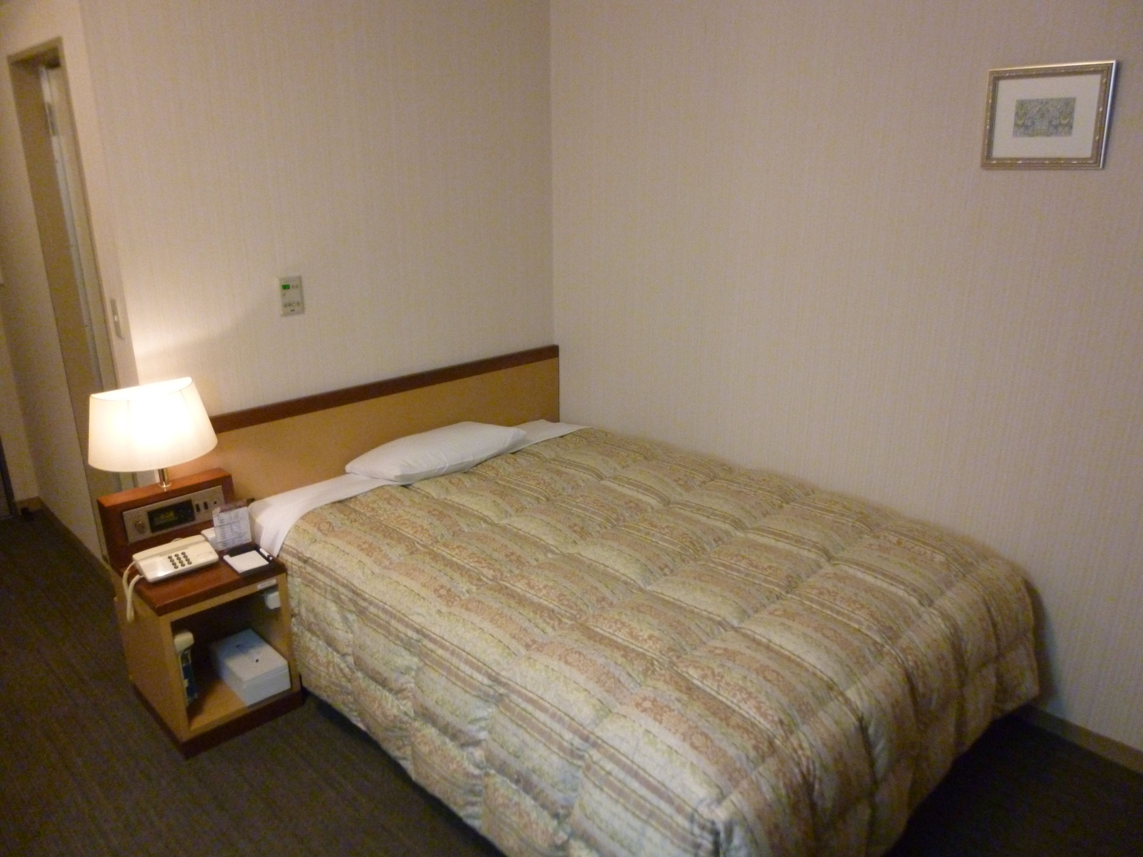 Una habitación de hotel con una cama, una mesita de noche con lámpara, un teléfono y un pequeño estante con una caja.