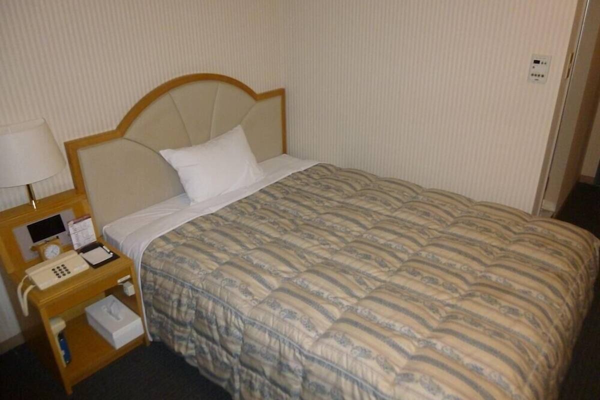 Een hotelkamer met een bed, een nachtkastje met een lamp, een telefoon en een klein tafeltje met een klok en een kalender.