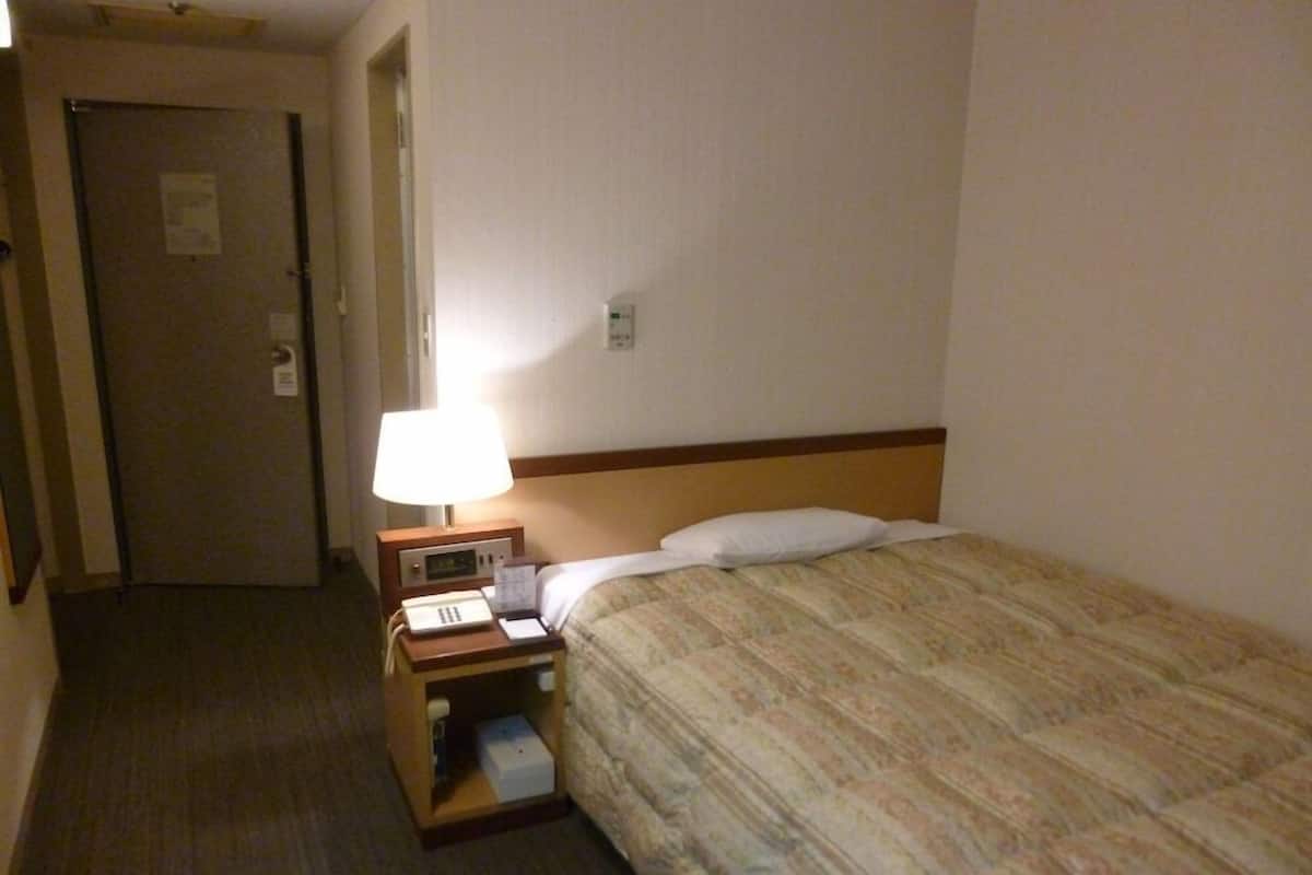 Een hotelkamer met een bed, een nachtkastje met lamp, een deur en een klein opbergmeubel.