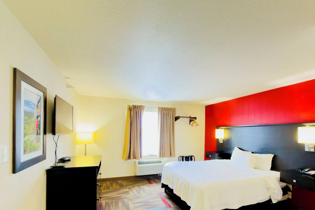 A hotel room with a bed, bedside tables, a desk, a chair, a lamp, a picture frame, a window with curtains, and a wall-mounted heater.