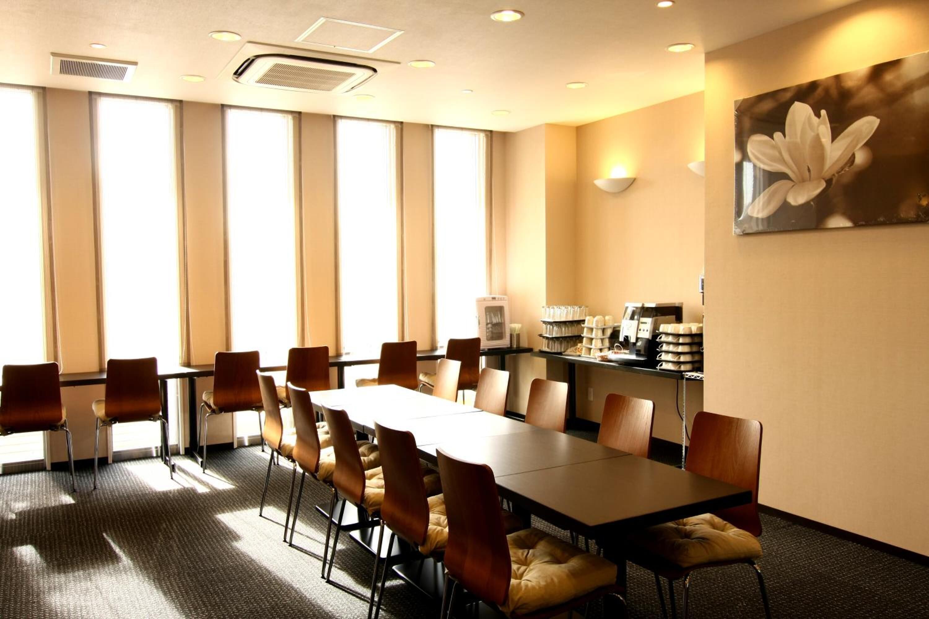 Un salón de conferencias con una larga mesa, varias sillas, una estación de café y ventanas amplias.