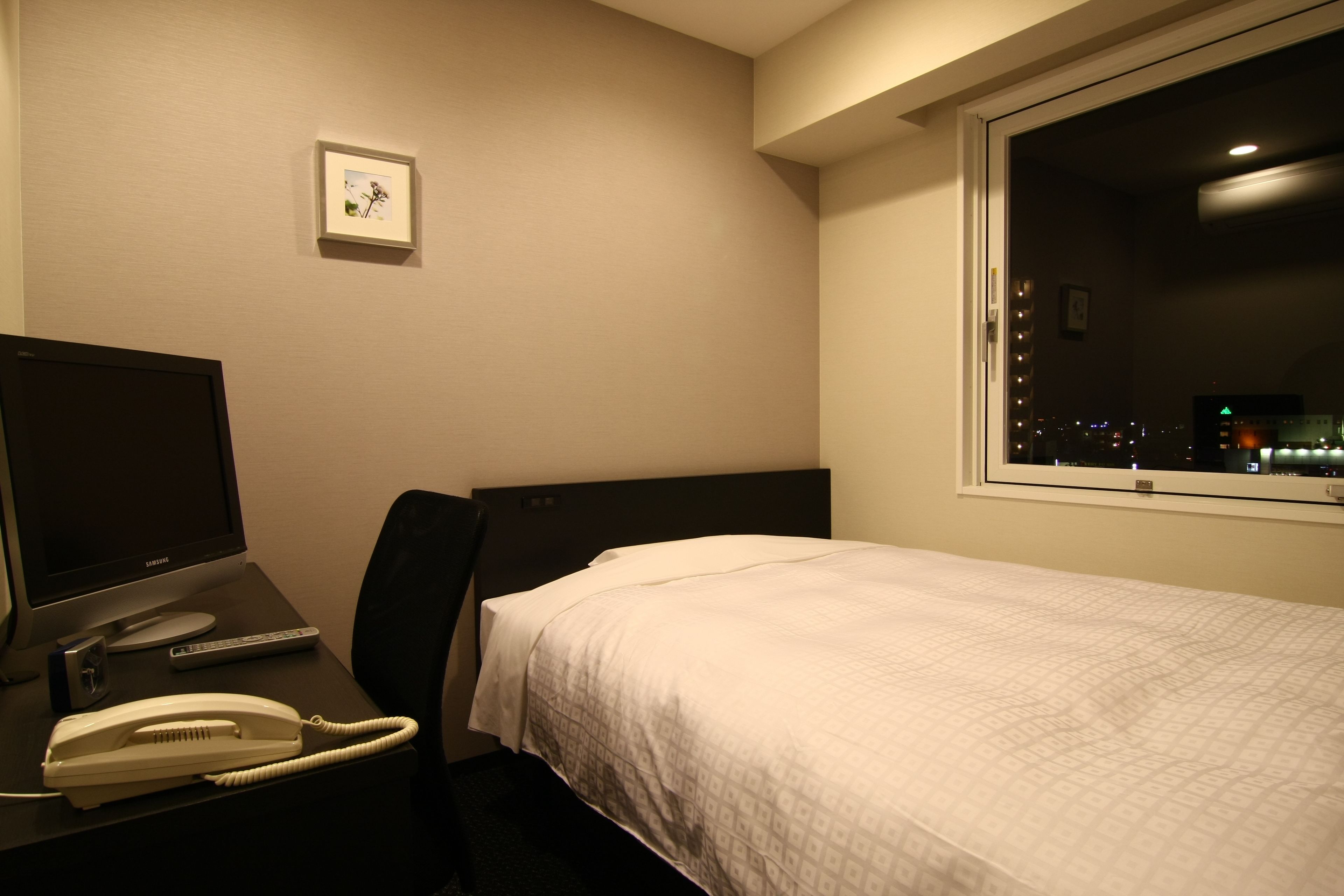 Ett hotellrum med en säng, ett skrivbord med en datorskärm, en telefon och ett fönster med utsikt över staden.
