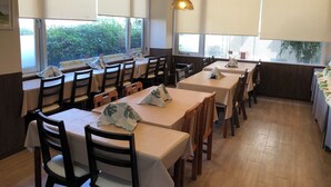 Um restaurante com várias mesas e cadeiras, amplas janelas com persianas e iluminação pendente.