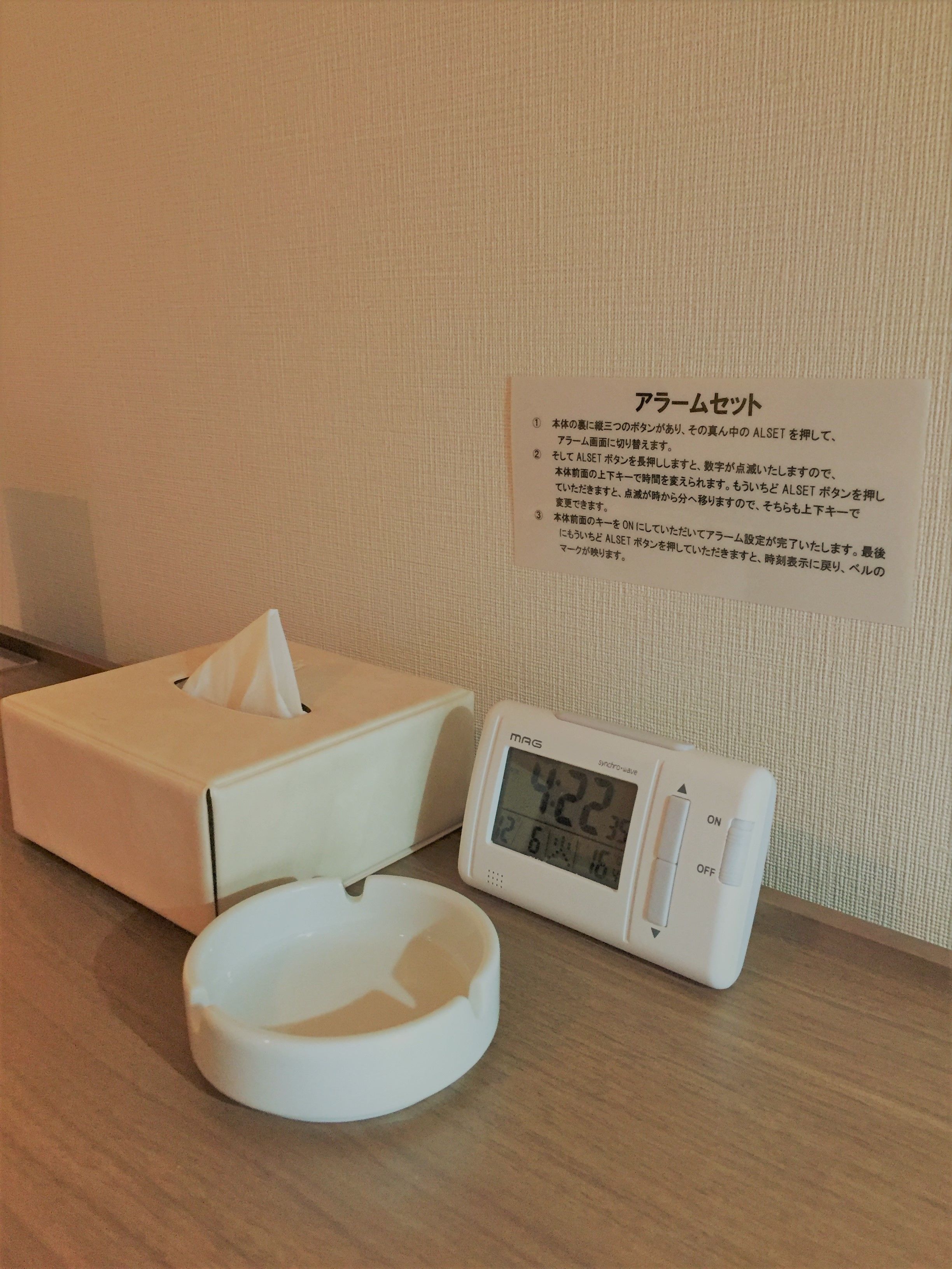 Una mesita de noche con un dispensador de pañuelos, un reloj digital que marca las 4:22 y un cartel con texto en japonés.
