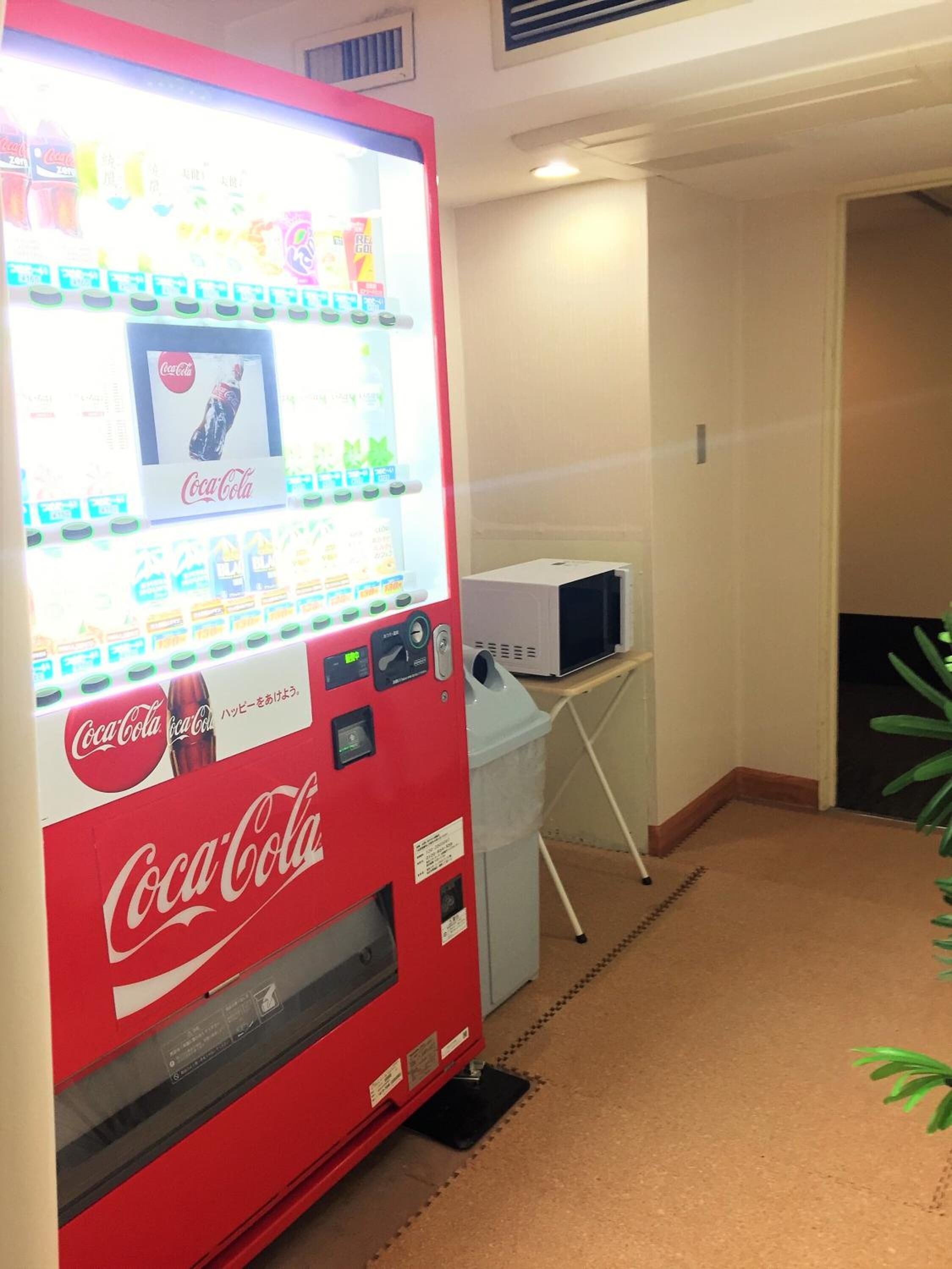 Máquina expendedora con diversas opciones de bebidas, incluyendo Coca-Cola, en una habitación con microondas y un cubo de basura.