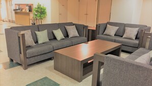 Ein modernes Wohnzimmer mit einer Eckcouch, einem Couchtisch und einem kleinen Beistelltisch. Im Hintergrund ist eine Rezeption mit einer Pflanze und einer Kaffeemaschine zu sehen.